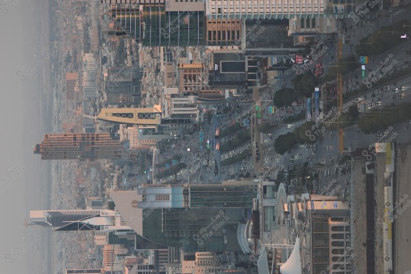 صورة تظهر منظرًا جويًا لمدينة حديثة مع العديد من المباني الشاهقة والشوارع المزدحمة بالسيارات. يمكن رؤية برج مميز يرتفع فوق بقية الهياكل, مع العمارة التي تجمع بين التصميم العصري والتقليدي. الشوارع تحيط بها المساحات الخضراء والأشجار التي تضفي جمالًا على المشهد الحضري.