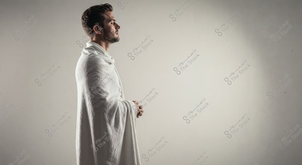 A side-profile portrait of a man wearing white clothing resembling an ihram, standing in a contemplative pose with his hands clasped. The background is plain white, adding a sense of calm and tranquility.