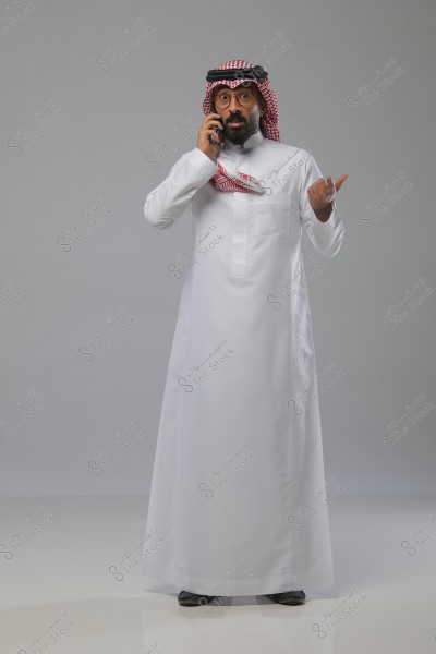 A portrait of a man wearing traditional Saudi attire, including a white thobe, red checkered headscarf (ghutra), and agal, while talking on a mobile phone and gesturing with his hand. The background is gray, and he is shown standing fully.