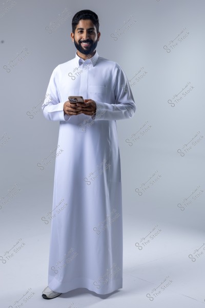 صورة لرجل يرتدي ثوباً أبيض تقليديًا ويحمل هاتفًا محمولًا في يديه, ويقف على خلفية بيضاء. الثوب يشير إلى زي عربي تقليدي شائع في دول الخليج مثل السعودية.