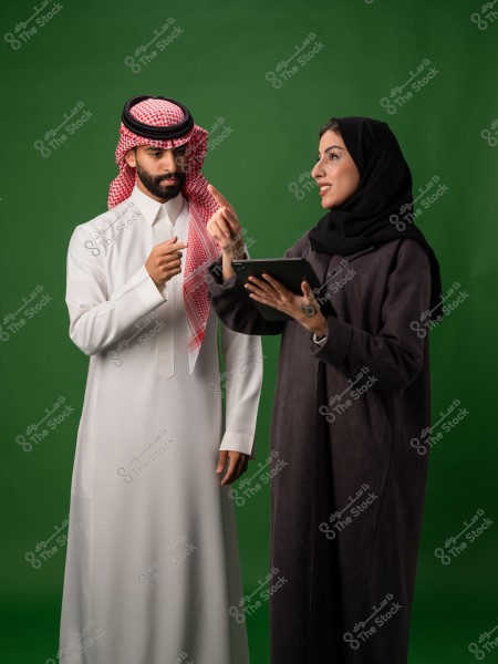 رجل يرتدي الزي السعودي التقليدي وامرأة محجبة يحملان جهازًا لوحيًا ويتبادلان النقاش على خلفية خضراء.