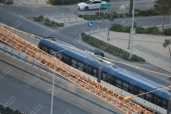 Aerial view of a modern metro train traveling on an elevated bridge in a city. The metro is blue with white stripes, surrounded by a bridge with orange supports. In the background, tree-lined streets and cars are visible near the metro, with a road sign indicating traffic directions.