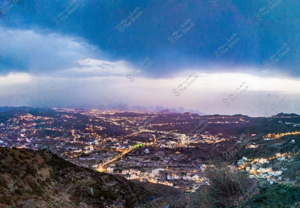 Panoramic view of a city illuminated at night, surrounded by hills and a dark sky. The city lights twinkle across the horizon, with rocky terrain framing the scene. Thick clouds cover part of the sky, suggesting an approaching storm.