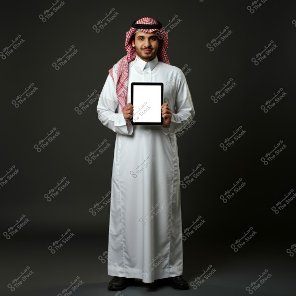 An image of a man wearing traditional white thobe and keffiyeh, holding a tablet with a blank screen, appearing to be from Saudi Arabia with a red-and-white checkered headscarf and agal. He stands against a dark background.