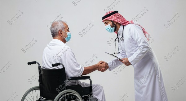 A doctor in a white coat, stethoscope, and face mask shakes hands with an elderly man sitting in a wheelchair, wearing white clothing and a face mask as well. The doctor is wearing a traditional Saudi headdress. The background is a simple white room.