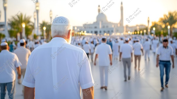 صورة لمجموعة كبيرة من الناس يسيرون في اتجاه مسجد كبير في الخلفية وقت الغروب. يرتدي الكثير من المشاركين ملابس بيضاء وقلنسوات. يتميز المسجد بمآذنه العالية وتصميمه المعماري المميز. الأرضية ممهدة وتحيط بها أشجار النخيل.