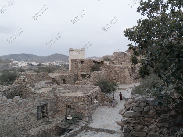 View of a traditional old village built from stone located in a mountainous area. The image shows stone buildings and a few people walking along the pathways between the houses. Trees are scattered throughout the scene, and hills and a group of modern city buildings are visible in the background.