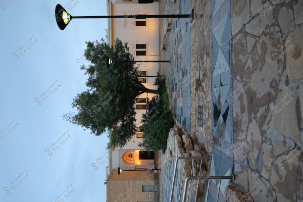 The image shows a courtyard in front of a traditional building with a white facade and ornate windows, adorned with illuminated outdoor lights. The building is surrounded by green trees and a pathway paved with stones in various colors. A street lamp is visible in the middle, indicating it is either dusk or dawn.