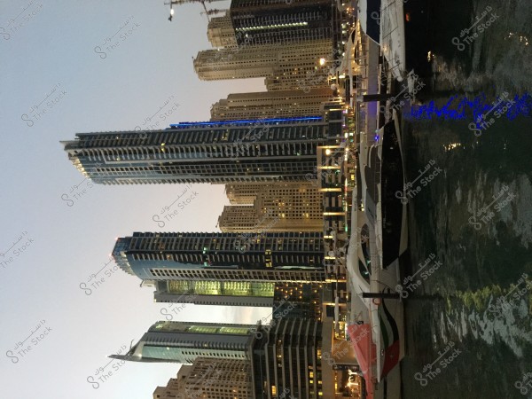 Tall skyscrapers alongside a waterfront in the Dubai Marina area, with several boats and yachts docked. The city’s night lighting is clearly visible, reflecting in the water with sparkling buildings.