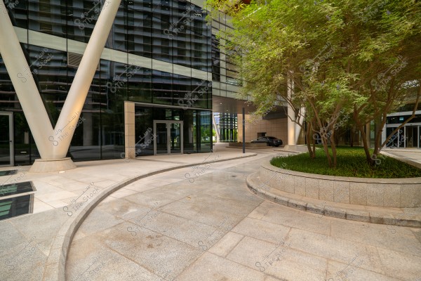 View of a modern building featuring a large glass facade and massive metal columns. The area is surrounded by marble flooring and a green tree in the right corner, giving an urban modern feel to the location.