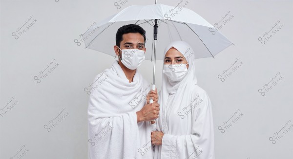 A portrait of a couple standing together under a white umbrella. The man is wearing traditional white Ihram garments, while the woman is wearing a white hijab and abaya. Both are wearing white medical masks. The background is plain white, providing focus on the couple and their religious attire.
