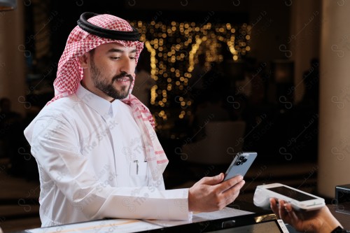 شاب سعودي في استقبال الفندق ، ويقوم بتعابير مختلفة باستخدام الجوال  ، ويقوم بدفع عبر جهاز الدفع الالكتروني