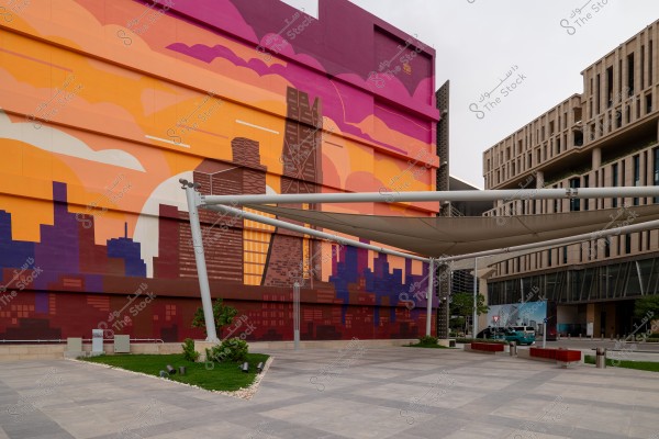 An image of a large wall adorned with a colorful mural depicting a cityscape in an artistic style with orange and pink skies. The image also shows a large tiled plaza with several small trees and a large white canopy. The neighboring building is modern in design and situated in an urban area.