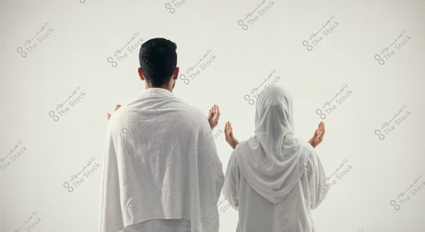 Image of a man and a woman from the back wearing white Ihram clothing, with their hands raised in prayer. The man is wearing a white wrap, while the woman is wearing a white headscarf. They are set against a simple white background.