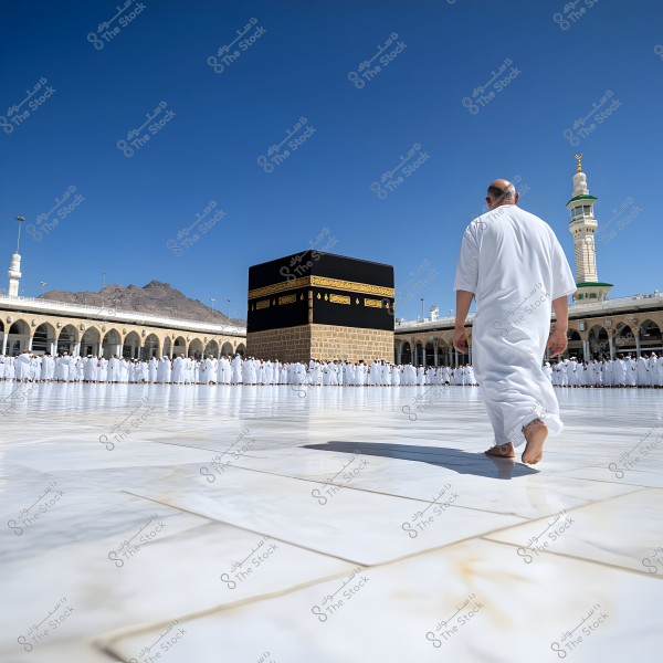 The image shows a person wearing a white Ihram walking towards the Kaaba in the Grand Mosque in Mecca. The weather is sunny with a clear blue sky, and worshippers are seen standing in a circular formation around the Kaaba. A white tower and minaret are visible in the background.