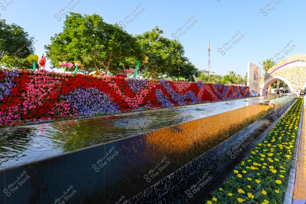 A colorful flower garden featuring a wall covered with red, purple, and pink flowers, with models of flowers and decorative structures on top. A water pathway runs along the right side, bordered by a path decorated with bright yellow flowers. Green trees provide shade to the garden, with a colorful flower arch visible in the background. The sky is bright and blue.