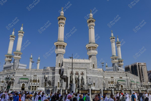 Image of the Great Mosque of Mecca in Saudi Arabia, featuring several tall white minarets adorned with gold detailing, surrounded by intricately designed marble walls. In the foreground, a group of people wearing various outfits, mostly white robes, suggesting a religious gathering, under a clear blue sky.