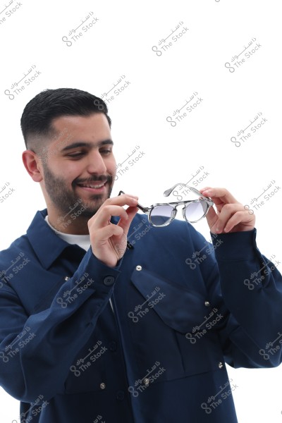 A portrait of a smiling man holding glasses in his hands. The man has a short beard and is wearing a blue shirt. The photo is taken against a white background, drawing attention to his features and the glasses he is holding.