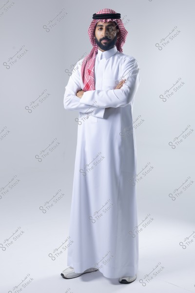 A portrait of a man standing with his arms crossed, wearing traditional Saudi attire consisting of a white thobe and a red and white checkered ghutra with a black agal. The background is white, adding a bright tone to the image.