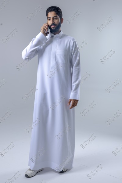 A portrait of a man standing in a studio wearing a traditional white thobe, holding a mobile phone to his ear. The background is light grey, and the man appears to be talking or listening attentively.