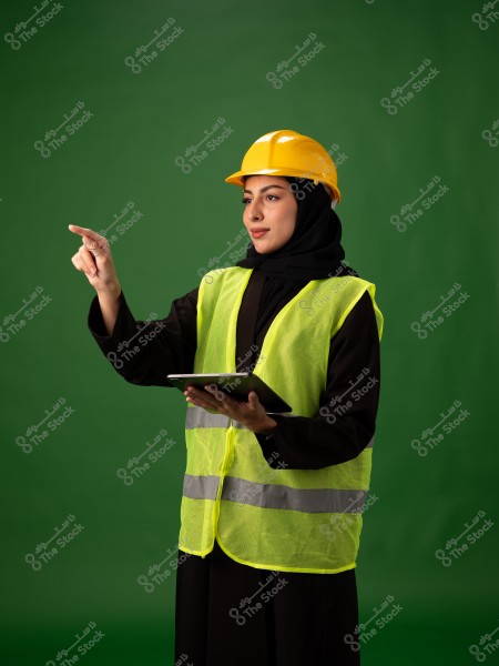 صورة لامرأة ترتدي حجابًا وملابس سوداء, مع خوذة بناء صفراء وسترة أمان صفراء لامعة. تحمل جهازًا لوحيًا وتشير بإصبعها, والخلفية خضراء. يبدو أنها تعمل في مجال الهندسة أو البناء.