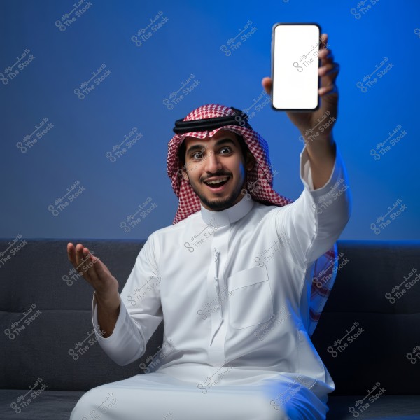 A man sitting on a sofa holds a smartphone in his hand raised up, with the phone’s screen appearing white and blank. He is wearing traditional Saudi attire, consisting of a white thobe, a red and white ghutra, and an agal. He smiles in an engaging manner, and the blue background adds a calm and appealing effect to the photo.