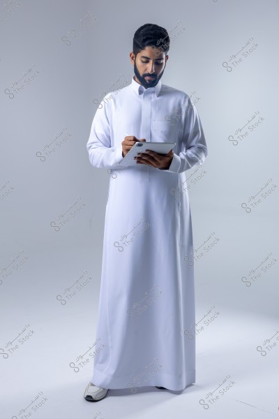 The image shows an individual wearing a white thobe, suggesting they are from a Gulf country. The person is standing and holding a tablet in their hands, using a stylus to write on it. The background is plain white, highlighting the person in the image.