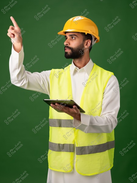 Image of a man wearing a traditional white shirt, a yellow safety vest, and a yellow hard hat, standing against a green background. He is holding a tablet in his left hand and pointing upwards with his right hand.
