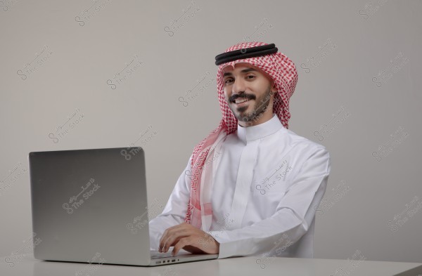 A portrait of a man sitting in front of a laptop. He is wearing traditional Saudi attire, which includes a white thobe and a red and white checkered ghutra with an agal. He is seated at a table in an indoor setting with a gray background.