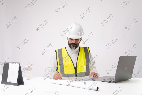 The image shows an engineer wearing a white safety helmet and a yellow reflective vest working at a desk. He is focused on reading blueprints on the table, with a laptop, a notepad, and an hourglass beside him. The engineer is wearing a traditional white thobe, suggesting he might be from the Gulf countries, possibly Saudi Arabia.