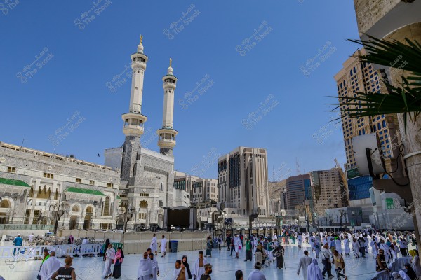 صورة لساحة المسجد الحرام في مكة المكرمة, السعودية. يظهر في المشهد المسجد المهيب بمئذناته الطويلة والجميلة, مع العديد من الأشخاص يرتدون ملابس بيضاء تقليدية للطواف حول الكعبة. يظهر في الخلفية عدة مبانٍ شاهقة والسماء زرقاء صافية.