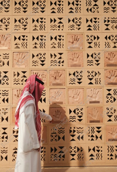 A man wearing traditional Saudi attire, including a thobe and a red-and-white checkered shemagh, walks beside a wall featuring repetitive geometric patterns and prominent handprints. The wall design includes triangular shapes and zigzags, with decorative details displayed in a series of squares.