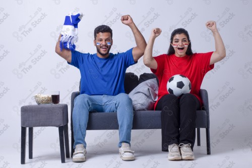 Portrait of a Saudi young man and woman watching a match and cheering their favorite teams cheerfully while having a snack on a blue background, the World Cup.