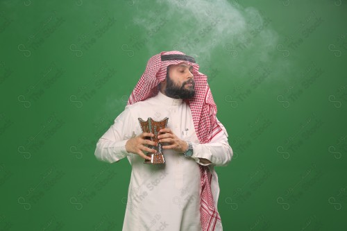 رجل سعودي يقف على خلفية خضراء يرتدي الثوب و الشماغ، يقوم بعمل حركات بيديه ، يبخر نفسه بالطيب و العود، احتفالات العيد