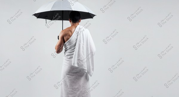 An image of a man from behind, holding a black and white umbrella. He is wearing two white cloths resembling the Ihram attire worn by pilgrims in Mecca. The background is completely white, which adds to the simplicity of the image and highlights the man in the center.