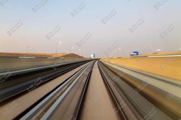 Image of a train moving swiftly on tracks, with blurred lines indicating speed. The sky is clear, suggesting early evening. Desert hills are visible in the background, and there are illuminated signs on either side of the track.