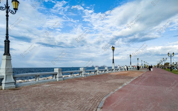 A long promenade facing the sea, lined with classic street lamps, with a cloudy sky and a partial rainbow visible. People are walking and strolling along the pathway with green spaces on the right and ships visible in the distance.