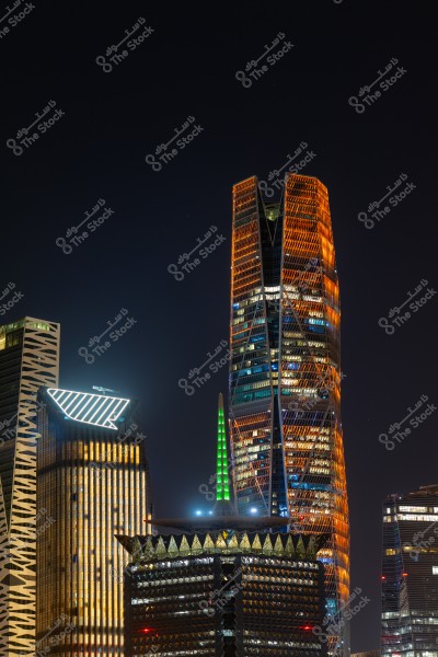 A night view of several skyscrapers in Riyadh, Saudi Arabia. The King Abdullah Financial District tower is lit in orange and blue, surrounded by other buildings with dazzling night lights. The black sky accentuates the bright lights of the structures.