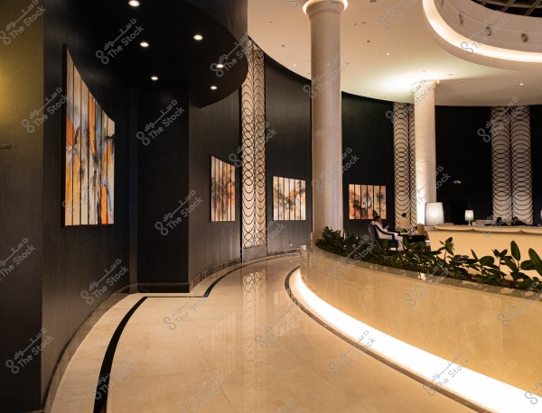 The image shows a modern reception area in a hotel or shopping mall, with dark walls adorned with illuminated abstract paintings. A curved path leads to the left with glossy floors and large white columns. Plants and botanical decorations adorn the area, with people sitting on sofas in a warmly lit seating area.