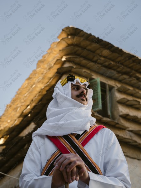 A snapshot of a Saudi man wearing traditional costume on the founding day, standing in front of a mud building and leaning on a stick, traditional costume, foundation day, ancient ancient buildings