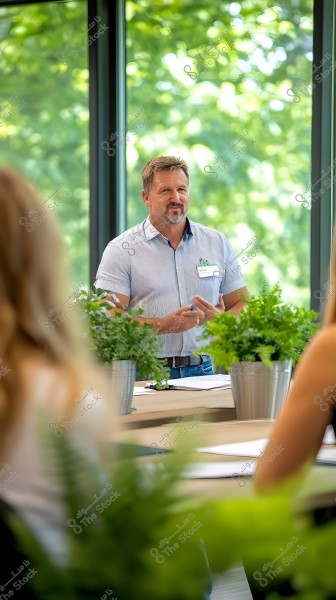 Image of a man wearing a short-sleeved striped shirt standing and giving a presentation in a meeting room filled with green plants. A table with papers and plant pots surrounds him. The background shows large windows overlooking a green natural landscape outside.