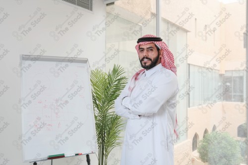 رجل سعودي يرتدي الزي السعودي التقليدي يقف مكتوف اليدين أثناء إجتماع في العمل نهارًا