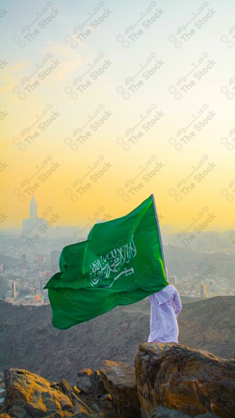 A snapshot of a Saudi man in traditional Saudi dress holding the Saudi flag over a mountain in the Makkah Al-Mukarramah region, the sky, the royal clock tower building in the Grand Mosque, buildings and landmarks, the Grand Mosque of Mecca.