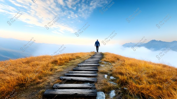 A man walks on a wooden pathway towards a mountain peak in a stunning natural landscape. Golden grassy plains stretch on either side of the path, with clouds enveloping the scene in the distance. Mountains appear on the horizon under a clear blue sky with some clouds.