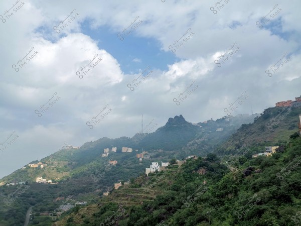 A scenic view of green mountains in a hilly area, with white and yellow buildings scattered on the mountainsides. The sky is cloudy with patches of blue visible. Communication antennas are present on the highest peak of the mountain. Dense vegetation covers the slopes.