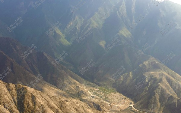 صورة جوية لمنظر طبيعي لجبال متعرجة, حيث تظهر التلال ذات الألوان البنية والظلال الطويلة. في الجزء السفلي من الصورة, يمكن رؤية طريق ضيق يتعرج بين الجبال, مع بعض المساحات الخضراء القليلة المتناثرة, مما يشير لوجود غطاء نباتي في الوادي.