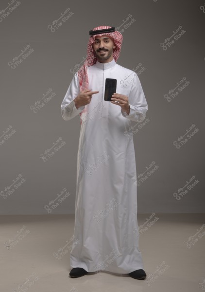 A portrait of a man standing in a studio wearing a white thobe, red ghutra, and black agal, indicating traditional Saudi attire. The man is smiling and pointing with his right hand at a smartphone he holds in his left hand.