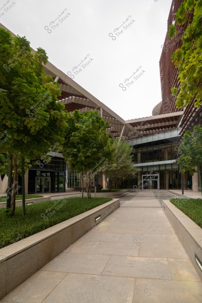 The image shows an outdoor courtyard of a modern building, surrounded by green trees and landscaping on both sides. The building features glass and metal facades, with a roof of contemporary architectural design. The sky is overcast, adding a serene feel to the atmosphere.
