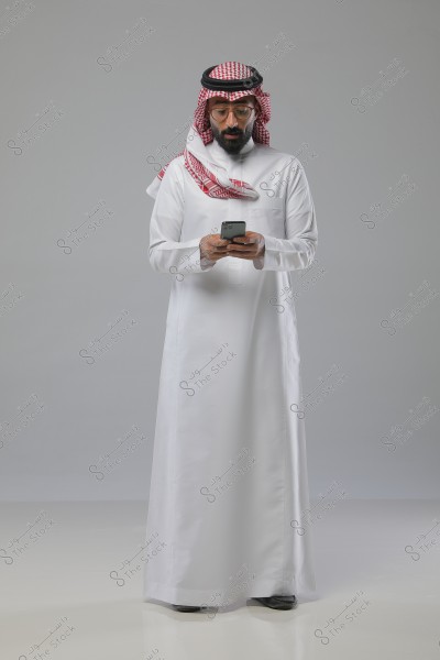 A man wearing a white thobe, red and white ghutra, and a black agal, appearing to be from the Gulf countries, holds a phone and looks at it. The background is plain gray.