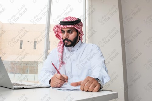 رجل سعودي يرتدي الزي السعودي التقليدي يعمل في مكتب بواجهة زجاجية ويستخدم قلم رصاص لتدوين الملاحظات